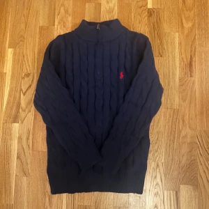 Mörkblå stickad half zip från Ralph Lauren - Mörkblå stickad tröja från Ralph Lauren med klassiskt kabelstickat mönster och röd broderad logga på bröstet. Tröjan har half zip, hög krage och långa ärmar. Perfekt för kyliga dagar och snygg till jeans eller chinos.