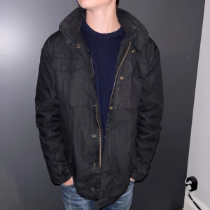 Svart parkajacka från Brandit - Svart parkajacka från Brandit med hög krage, flera fickor och dragkedja med knappar. Jackan har en klassisk, rak passform och är tillverkad i slitstark polyester. Perfekt för dig som vill ha en stilren och funktionell jacka till vardags. Skriv vid fler bilder eller frågor!