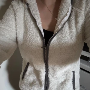 Vit teddyjacka med dragkedja - Mysig vit teddyjacka med fluffig känsla och hel dragkedja framtill. Jackan har två fickor med mörka detaljer och en hög krage. Perfekt för dig som vill ha en varm och stilren look under kyliga dagar. I storlek M men är mer som en XS/S! 