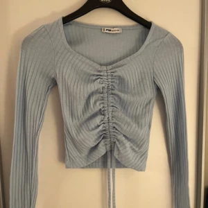 Ljusblå ribbad topp med snörning - Supersnygg ljusblå långärmad topp med ribbat tyg och snörning framtill som ger en cool rynkad effekt. Toppen har v-ringning och croppad passform, perfekt att styla med jeans eller kjol. Materialet är mjukt och stretchigt för extra komfort.