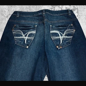 Evisu wide jeans med broderade fickor XL - Mörkblå jeans från Evisu i storlek XL med breda ben och ikoniska broderade bakfickor i vitt. Klassisk femficksmodell med kontrastsömmar och slitningar på benen. Perfekta för dig som gillar statement-plagg och streetwear.