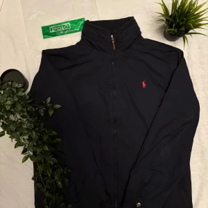 Svart vindjacka från Polo Ralph Lauren - Svart vindjacka från Polo Ralph Lauren med klassisk ståkrage och dragkedja framtill. Jackan har röd broderad logga på bröstet och är tillverkad i polyester. Långärmad modell med knappdetaljer vid ärmslut och enkel, stilren design.
