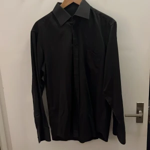 Svart skjorta från Shirts by Claudio - Säljer en svart skjorta från Shirts by Claudio med klassisk krage och lång ärm. Skjortan har en bröstficka och knäppning framtill. Snygg och stilren modell i slät bomull, perfekt för dig som gillar en clean look.