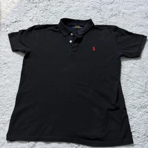 Svart pikétröja från Polo Ralph Lauren - Klassisk svart pikétröja från Polo Ralph Lauren med korta ärmar och röd broderad logga på bröstet. Tröjan har krage, knäppning med två knappar och är gjord i mjuk bomull. Perfekt för en clean och stilren look.