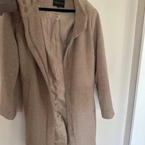 Beige kappa från STOCKH LM, stl 38 - Snygg beige kappa från STOCKH LM i klassisk rak modell. Jackan har dragkedja framtill, hög krage med knappar och två fickor. Tillverkad i mjukt ullmaterial med slät insida. Perfekt för dig som vill ha en stilren och tidlös look.
