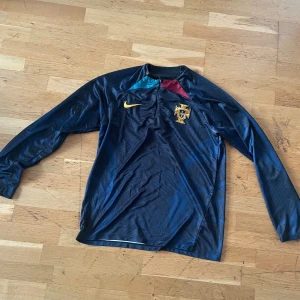 Portugal långärmad fotbollströja Nike - Snygg långärmad fotbollströja från Nike med Portugals landslagsemblem på bröstet. Tröjan är mörkblå med detaljer i gult, rött och turkos vid halsen samt gul Nike-logga. Tillverkad i lätt och ventilerande material, perfekt för träning eller match.