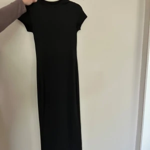 Svart långklänning med kort ärm - En stilren svart långklänning med rund hals och korta ärmar. Klänningen har en rak och enkel passform som ger en clean look. Perfekt för dig som gillar minimalistisk stil och vill ha något som funkar till många olika tillfällen.