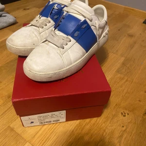 Valentino skor  - Säljer ett par Valentino Garavani sneakers i vitt skinn med bred blå läderdetalj över snörningen och ikoniska nitar runt hälen. Klassisk rund tå, platt sula och vita skosnören.  