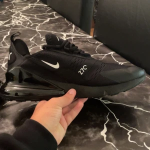 Nike Air Max 270 svarta sneakers - Svarta Nike Air Max 270 sneakers med vit logga och detaljer. Skorna har en stor synlig Air-enhet i hälen, mesh-ovandel och snörning. Snygg och sportig design som passar perfekt till streetwear. Materialet är syntet och mesh för bra andningsförmåga.