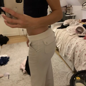 Vita Juicy byxor  - Snygga beiga mjukisbyxor med bred midja och dragsko framtill. Byxorna har raka ben, stora fickor med lock och knappdetaljer där bak. Perfekta för en chill och avslappnad stil. Lite så defekter där nere