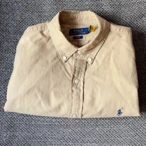 Ralph Lauren Skjorta Beige - Snygg beige skjorta från Polo Ralph Lauren i slim fit. Klassisk button-down krage, långärmad och med blå broderad logga på bröstet. Storlek M, aldrig använd, har bara legat i garderoben i ett år. Skick 10/10❗️ Tillverkad i mjuk bomull och har vita knappar framtill. Perfekt för dig som gillar stilrena och tidlösa plagg. Nypris 1.799kr på J:B:S!