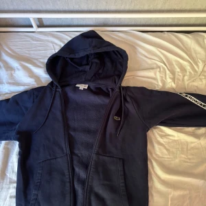 Mörkblå Lacoste hoodie med dragkedja - Säljer en mörkblå hoodie från Lacoste i klassisk passform. Priset är diskuterbart, möts i Stockholm. Tröjan har huva med snörning, dragkedja framtill och vita Lacoste-loggor längs ärmarna. Mjuk insida och två fickor framtill. Perfekt för chill dagar eller när du vill ha en sportig look.