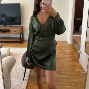 Olivgrön omlottklänning från Zara - Snygg olivgrön kort omlottklänning från Zara i silkes material. Klänningen har långa ärmar med breda manschetter, v-ringning och markerad midja. Perfekt för dig som gillar en stilren och trendig look.