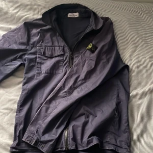 Stone Island vindjacka i mörkblått - Snygg mörkblå vindjacka från Stone Island med klassisk logotyp på ärmen. Jackan har dragkedja framtill, bröstficka med lock och lång ärm. Perfekt för dig som gillar streetwear och vill ha en stilren look. Äkta stone island jacka priset är inte hugget i sten 