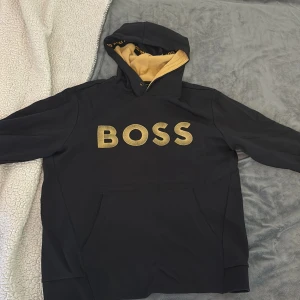 Svart hoodie från BOSS med guldtryck - Svart hoodie från BOSS med stort guldigt BOSS-tryck på bröstet och kontrasterande beige insida på huvan. Klassisk känguruficka framtill och huva med snörning. Perfekt för dig som vill sticka ut med en snygg och bekväm tröja.