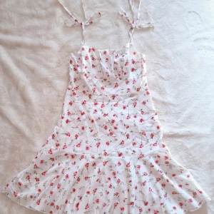 Vit blommig kortklänning med knytband - Supersöt vit kortklänning med röda och rosa blommor, smala axelband med knyt och lätt utsvängd kjol. Perfekt för varma dagar och har en luftig, feminin känsla. Materialet känns lätt och svalt mot huden.