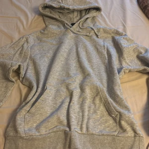 Grå hoodie med huva och ficka - En klassisk ljusgrå hoodie med huva och snörning. Tröjan har en stor magficka och ribbade muddar vid ärmslut och nederkant. Perfekt för chill dagar eller när du vill ha något bekvämt och stilrent.