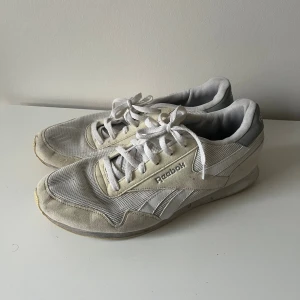 Vita Reebok sneakers med mesh och mocka - Klassiska vita Reebok sneakers med lågt skaft, snörning och platt sula. Ovandelen är i mesh och mocka med grå detaljer och Reebok-logga på sidan och hälen.