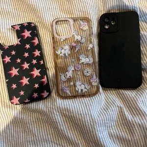 iPhone 12 Skal, 3-pack - Tre skal till iPhone 12: ett svart med rosa stjärnor (skadat hörn), ett genomskinligt med enhörningar och ett helsvart skal. Passar iPhone 12, skyddar mot repor och stötar. Skalen är använda, ett har tydligt slitage, de andra i gott skick.