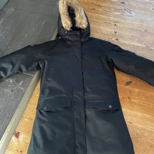 Svart dunjacka med fuskpäls från Everest - Svart lång dunjacka från Everest med huva och fuskpäls. Jackan har dragkedja, tryckknappar, två stora fickor med lock och två snedställda bröstfickor. Insidan är quiltad för extra värme och komfort. 