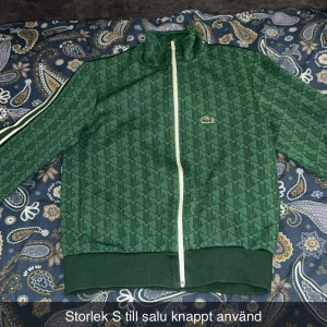 Grön mönstrad ziptröja från Lacoste - Säljer en grön ziptröja från Lacoste med diskret geometriskt mönster och vit dragkedja. Tröjan har ribbade muddar, hög krage och klassisk Lacoste-logga på bröstet. Perfekt för en sportig och trendig look.