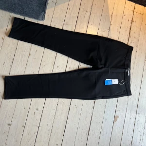 Svarta kostymbyxor från Samsøe Samsøe - Snygga svarta kostymbyxor från Samsøe Samsøe, modell Frankie Pants. Byxorna har en rak passform med klassisk midja och stängs med knapp och dragkedja. Perfekta till en clean och stilren look. Materialet är mjukt och följsamt för en bekväm känsla.