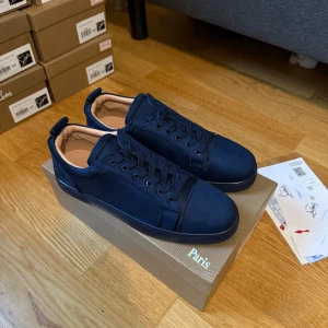 Mörkblå sneakers från Christian Louboutin storlek 44 - Snygga mörkblå sneakers från Christian Louboutin med lyxig mocka och klassisk röd sula. Skorna har rund tå, snörning och broderad logga på hälen. Insidan är beige och ger en exklusiv känsla. Perfekt för dig som gillar stilrena detaljer och vill sticka ut.