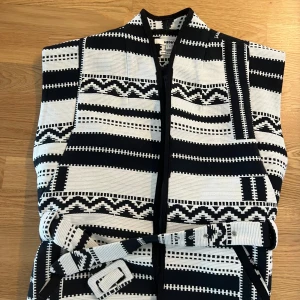 Svartvit mönstrad väst med bälte - Snygg svartvit väst från Koton med grafiskt randigt mönster och tillhörande bälte i midjan. Västen har en rak passform och är ärmlös, perfekt för att lyfta vilken outfit som helst med sin unika design.