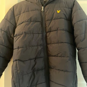 Svart pufferjacka från Lyle & Scott - Svart pufferjacka från Lyle & Scott med gul logga på bröstet. Jackan har huva, dragkedja framtill och quiltad design. Perfekt för kyliga dagar och riktigt skön att bära. Passar dig som gillar en enkel men snygg stil. Jackan har ett litet hål på ärmen, därav billig. Annars i jättebra skick. 