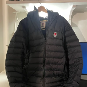 Svart dunjacka Fjällräven Expedition M - Säljer en svart Fjällräven Expedition Series dunjacka i storlek M. Jackan har klassisk pufferdesign med huva, dragkedja och ikoniskt rött Fjällräven-märke på bröstet. Perfekt för kalla vinterdagar och inspirerad av originalet från 1974.