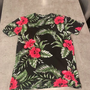 Blommig t-shirt från SMOG, svart/grön - Snygg t-shirt från SMOG i regular fit med svart bas och stora röda blommor samt gröna blad. Perfekt för dig som vill sticka ut med ett tropiskt mönster. Kortärmad och skön att bära under varma dagar.