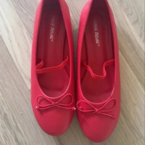 Röda ballerinaskor med rosett - Snygga röda ballerinaskor med rund tå och dekorativ rosett framtill. Skorna har en enkel rem över foten för extra stil och sitter bekvämt på foten. Perfekta för dig som vill ha en färgklick till outfiten.