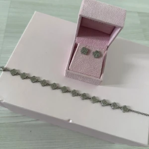 Silverfärgat armband & örhängen set - Snyggt set med armband och örhängen i silverfärg. Båda har små fyrklöverformade hängen täckta av glittrande stenar som ger en lyxig känsla. Perfekt för dig som gillar eleganta detaljer och vill sticka ut lite extra.