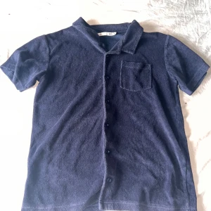 Mörkblå frottéskjorta från 157 - Mjuk och chill mörkblå skjorta från 157 i frotté och mocka. Kortärmad med klassisk krage, knappar hela vägen och en bröstficka. Perfekt för dig som gillar avslappnad stil och vill ha något annorlunda i garderoben.