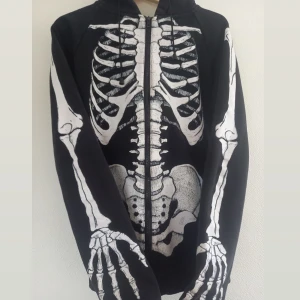 Svart hoodie med skelettprint - Cool svart hoodie med helskalet skelettmönster i vitt både fram och bak. Tröjan har huva , dragkedja och långa ärmar med detaljerade benmotiv. Perfekt inför Halloween. Djur finns i hushållet! 