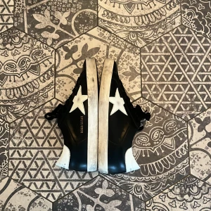 Golden Goose superstar - Svarta sneakers från Golden Goose med vit stjärna på sidan och vit häl. Skorna har svarta skosnören, vit sula med slitna detaljer. Tillverkade i skinn och har en klassisk låg profil. Denna sko kommer vara perfekt under vinterns eftersom den är svart och inte blir så smutsig. Köpte dessa skor från en kompis.