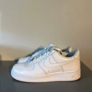Nike Air Force 1 vita sneakers - Klassiska Nike Air Force 1 sneakers i helvitt skinn med perforerad tå, rund tå och platt sula. Snygg och tidlös modell med snörning och diskret Nike-logga på sidan. Perfekta för dig som vill ha en clean och stilren look.