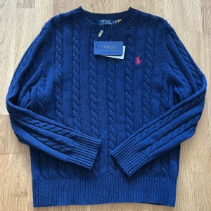 Marinblå kabelstickad tröja Polo Ralph Lauren - Kabelstickad marinblå tröja från Polo Ralph Lauren med klassisk rund halsringning och röd broderad logga på bröstet. Tröjan har långa ärmar och ribbade muddar för en snygg passform. Perfekt för dig som gillar stilren och tidlös design. Helt nytt, Fraktar inom 24 timmar. 