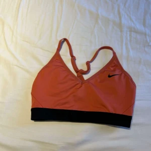 Nike sporttopp i orange och svart - Sportig och snygg sporttopp från Nike i orange med svart resår nedtill. Toppen har smala axelband som möts i en knut baktill och en diskret Nike-logga framtill. Perfekt för träning och gjord i ett stretchigt syntetmaterial som andas.