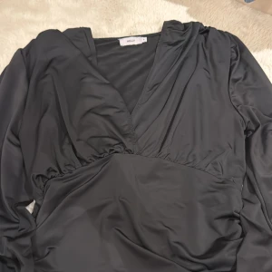 Svart topp från Nelly - Snygg svart topp från Nelly med långa ärmar och markerad midja. Toppen har v-ringning och är tillverkad i ett glansigt, mjukt material som ger en elegant känsla. Perfekt för dig som gillar stilrena och trendiga plagg.