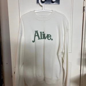 Vit sweatshirt med tryck 'Alive.' - Vit sweatshirt med grönt tryck 'Alive.' på bröstet. Tröjan har rund halsringning och långa ärmar. Perfekt för dig som gillar enkel och clean stil med ett statement. Mjuk och skön, passar till jeans eller mjukisbyxor.