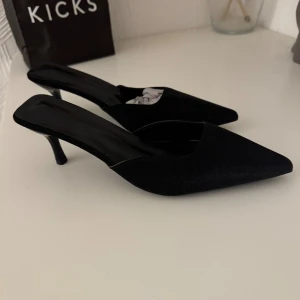 Svarta pumps med spetsig tå - Stilrena svarta pumps med spetsig tå och låg klack. Skorna har öppen häl och en elegant, minimalistisk design. Materialet är slätt och ger en lyxig känsla, perfekt för dig som gillar en klassisk och trendig look.