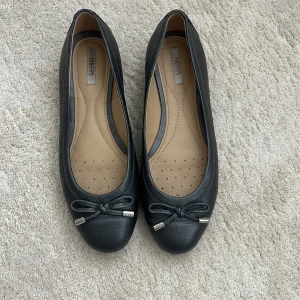 Svarta ballerinaskor  - Snygga svarta ballerinaskor i skinn med rund tå och dekorativ rosett framtill. Skorna har en beige innersula med små perforeringar för extra komfort. Perfekta för dig som vill ha en klassisk och stilren look.