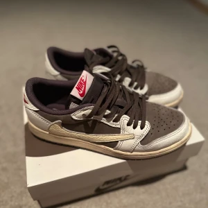 Nike Air Jordan 1 Low Travis Scott - Nike Air Jordan 1 Low Travis Scott med bruna och vita paneler i skinn och mocka, ikonisk omvänd Swoosh i beige, svarta snören och röd Nike-logga på tungan. Låg modell med perforerad tå och vintage-inspirerad sula. Perfekt för dig som vill sticka ut med hypade sneakers.