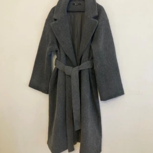 Grå kappa med bälte från Gina tricot  - Stilren grå kappa från Gina tricot  med klassisk krage och knytskärp i midjan. Kappan är lång, har raka ärmar.Perfekt för dig som vill ha en enkel men snygg look under höst och vinter.