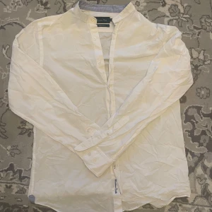 Vit henley-skjorta från Massimo Dutti - Säljer en stilren vit henley-skjorta från Massimo Dutti i casual fit. Skjortan har lång ärm, knappar framtill och en klassisk ståkrage. Perfekt att ha under din skjorta eller kavajen! Inga defekter, aldrig använd, som ny!