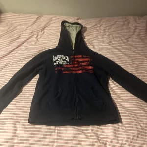 Mörkblå hoodie från Ralph Lauren L - Mörkblå hoodie från Ralph Lauren med dragkedja, huva och tryck i vitt och rött framtill. Snygg streetstil med text och grafiskt mönster på bröstet. Mjuk insida och två fickor framtill. Perfekt för dig som gillar klassiska märken och sportig look.