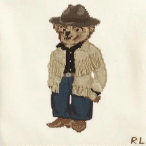 Vit stickad tröja med cowboy-motiv RL - Vit stickad tröja från Ralph Lauren med broderad ikonisk nalle i cowboy-outfit framtill. Tröjan har en klassisk passform och RL-broderi i hörnet. Perfekt för dig som gillar unika detaljer och coola motiv.
