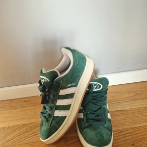 Adidas Campus gröna sneakers mocka - Gröna Adidas Campus sneakers i mocka med klassiska vita ränder på sidorna och vit sula. Skorna har rund tå, snörning och Adidas logga på plösen. Perfekta för dig som gillar retrostil och vill sticka ut med en snygg färg på fötterna.