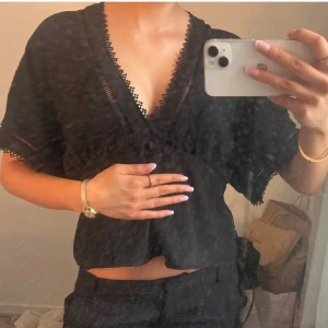 Svart blus med spetsdetaljer - Säljer en svart blus med djup v-ringning och fina spetsdetaljer längs halsringningen och ärmsluten. Blusen har korta, vida ärmar och en lös passform som ger en avslappnad vibe. Perfekt att styla med höga jeans eller kjol.